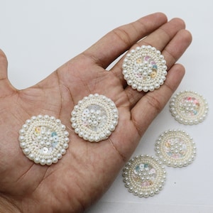 May include: Six white pearl and sequin embellishments. Each embellishment is a circle with a center of sequins and a border of white pearls.
