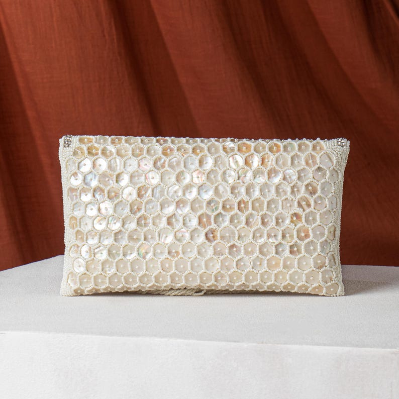 May include: A rectangular, off-white clutch with a honeycomb pattern of iridescent, shell-like pieces. The edges are trimmed with small, white beads. The clutch is set against a white surface and a rust-colored backdrop.