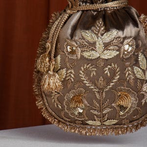 Könnte beinhalten: Ein brauner Satin-Beutel mit aufwendiger Goldstickerei. Das florale Design umfasst Blätter, Blüten und Pailletten. Goldene Quasten und Fransen säumen die Ränder und verleihen diesem Accessoire Eleganz.