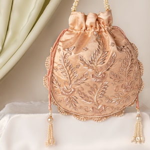 May include: A peach-colored drawstring bag with a floral design embroidered in gold thread. The bag has a pearl-trimmed border and a pearl tassel.