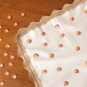 May include: A white fabric table runner with a gold scalloped border and a pattern of orange and red floral embellishments.