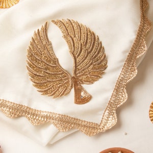 Puede incluir: Un cuadrado de tela blanca con un diseño bordado en oro de un par de alas. La tela tiene un borde festoneado.