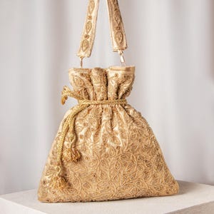 May include: A gold-colored drawstring bag with intricate embroidery and beaded details. The bag features a matching strap and a tassel. The design includes floral patterns and a drawstring closure. The bag is displayed on a white surface.
