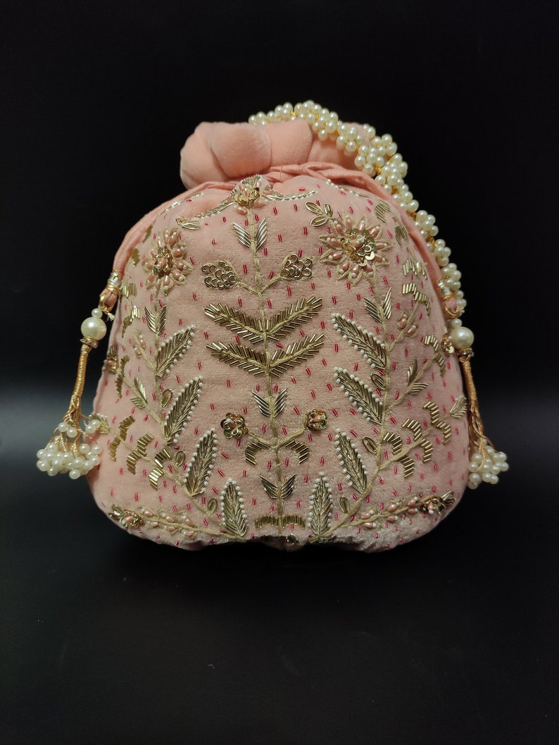 Puede incluir: Una bolsa de cord&oacute;n de terciopelo rosa con adornos dorados y de perlas. La bolsa est&aacute; decorada con un patr&oacute;n floral y tiene un borla de cuentas.
