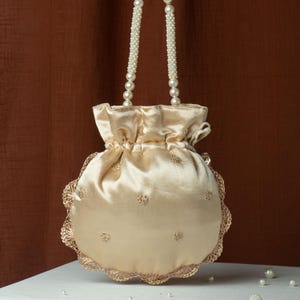 May include: A cream-colored satin drawstring bag with a scalloped lace trim. The bag features small embroidered floral details and pearl beaded handles. The background is a solid brown.