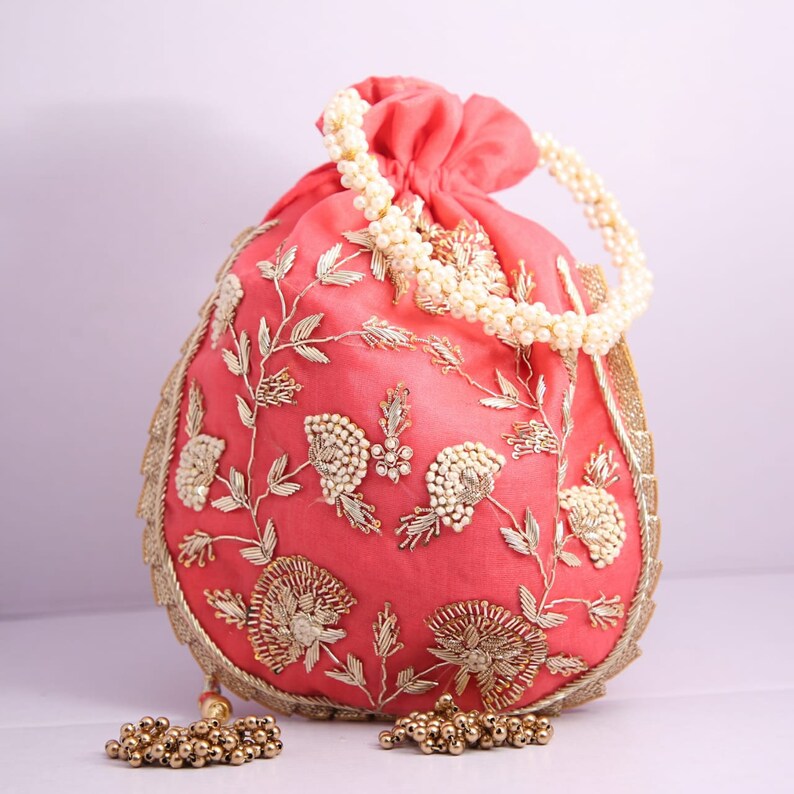 May include: A pink drawstring bag with gold and white embroidery and a pearl handle. The bag is decorated with floral patterns and has a tassel.