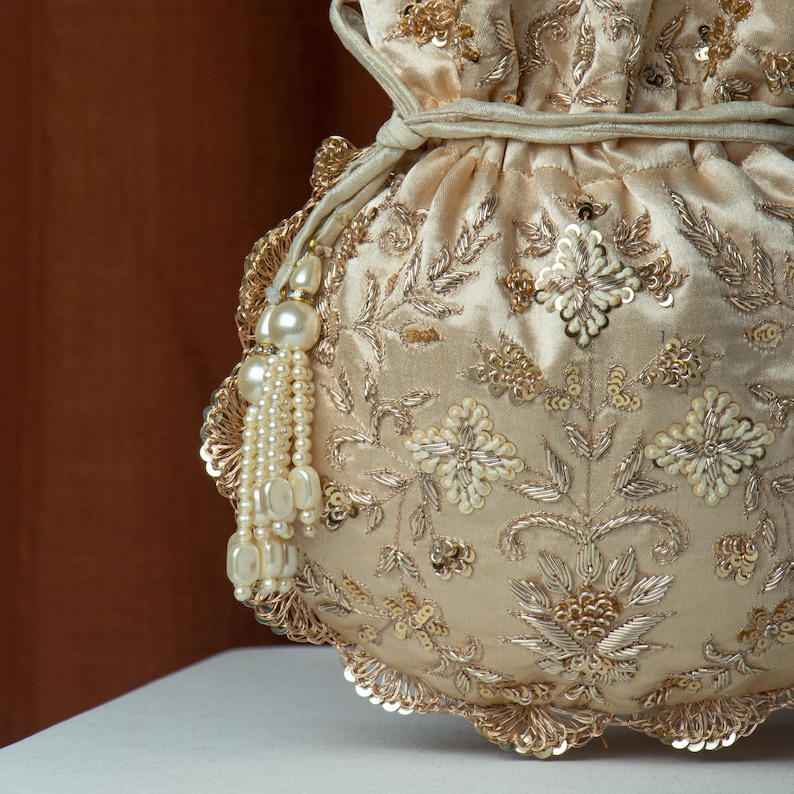 May include: A cream-colored, drawstring pouch with intricate gold embroidery and sequin details. The bag features floral patterns and a scalloped edge, with a pearl tassel. The pouch is made of a soft, satin-like material.