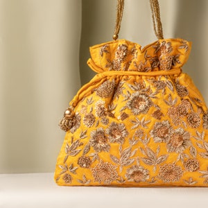 Puede incluir: Una bolsa de cordón amarillo con bordados dorados y lentejuelas. La bolsa tiene un patrón floral y un borla.