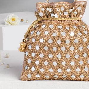 Puede incluir: Un bolso dorado con cordón y un patrón geométrico de pequeños espejos y abalorios dorados. El bolso tiene una borla y un cierre de cordón dorado. El bolso se utiliza probablemente para llevar objetos pequeños.
