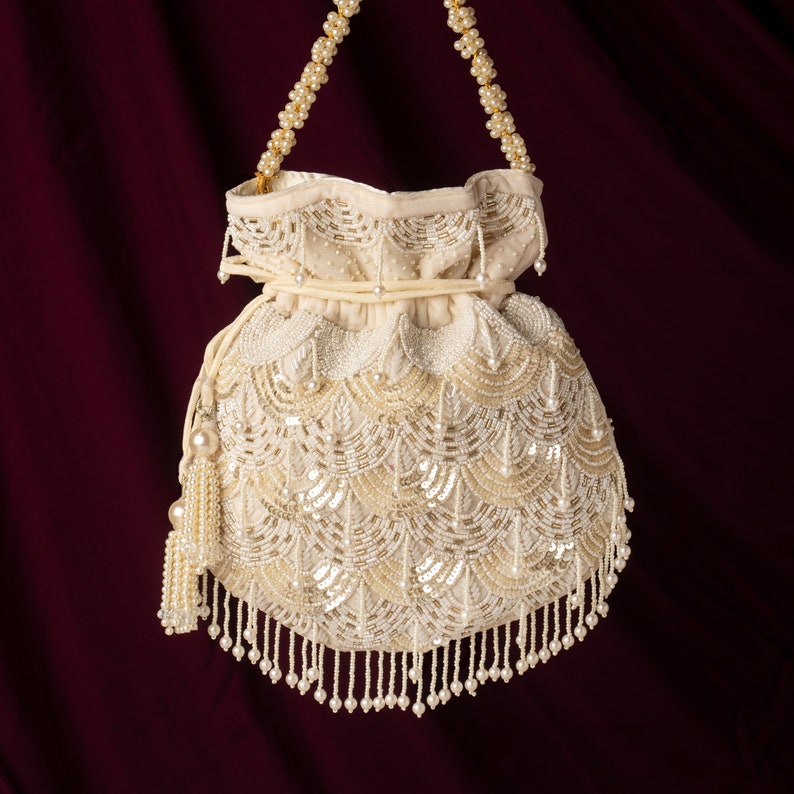 May include: A white beaded drawstring bag with a pearl chain strap and fringe. The bag is decorated with a pattern of white and gold beads.