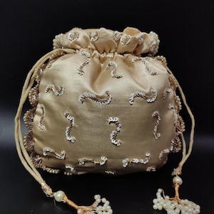 May include: A champagne-colored satin pouch bag with a drawstring closure. The bag is adorned with white sequins and gold embroidery. The edges are trimmed with gold lace and the drawstrings end in beaded tassels. The background is black.