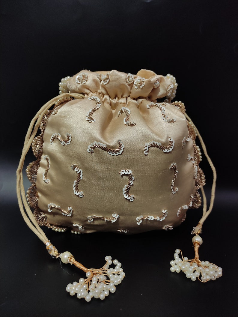 May include: A champagne-colored satin pouch bag with a drawstring closure. The bag is adorned with white sequins and gold embroidery. The edges are trimmed with gold lace and the drawstrings end in beaded tassels. The background is black.