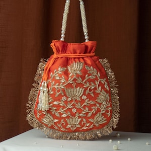 May include: An orange drawstring bag with gold floral embroidery. The bag has a beaded handle and a fringe of gold sequins. A tassel of pearls hangs from the side. The bag is sitting on a white surface.