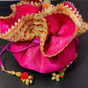 May include: A pink drawstring bag with gold and orange trim. The bag has a tassel with gold beads and a red, orange, and yellow pom-pom.