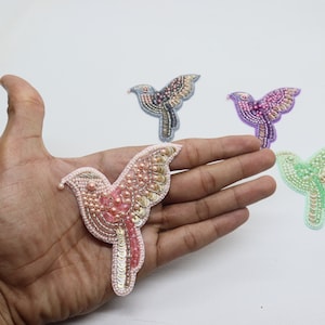 Puede incluir: Cuatro aplicaciones de pájaros con cuentas en diferentes colores: rosa, morado, verde y gris. Las aplicaciones están hechas de cuentas y lentejuelas y tienen un diseño detallado.