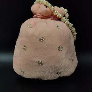 Puede incluir: Una bolsa de cord&oacute;n de terciopelo rosa con un asa de cadena de perlas y adornos dorados. La bolsa est&aacute; decorada con peque&ntilde;as flores brillantes.