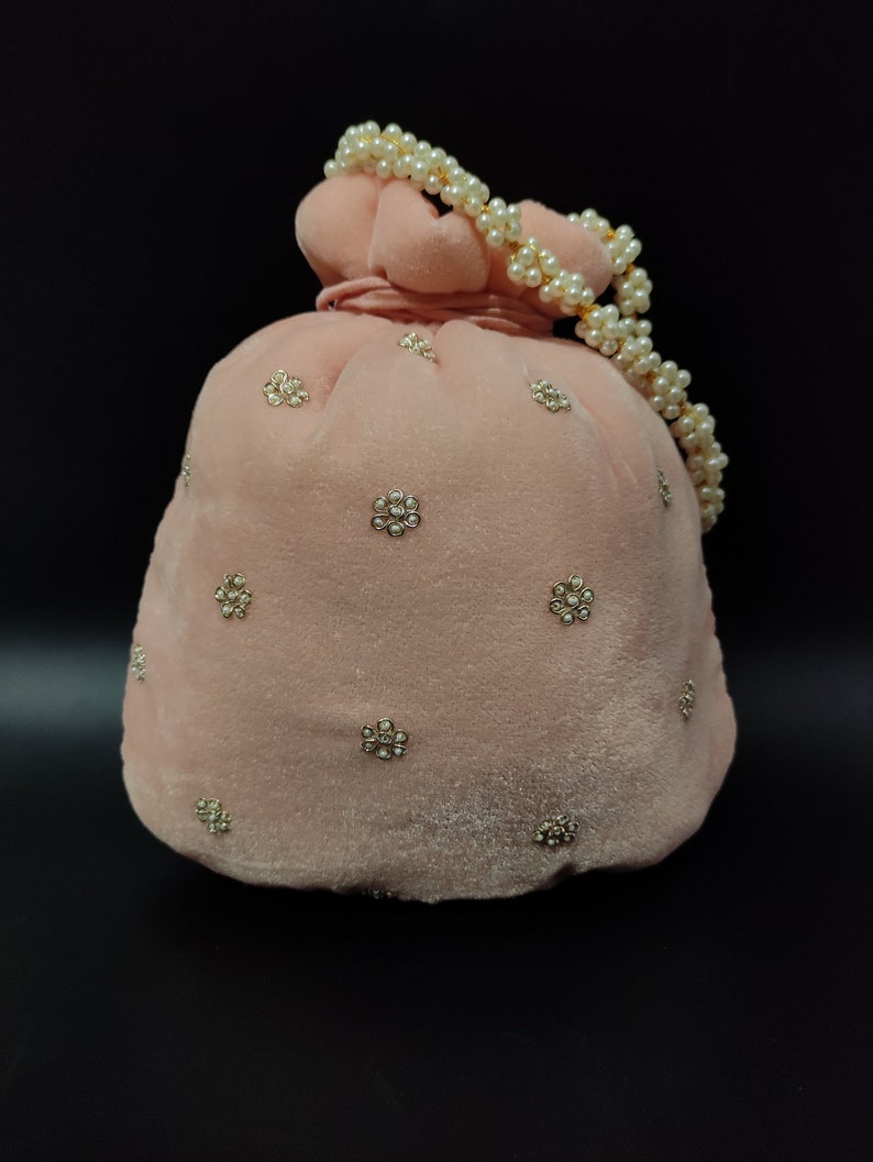 Puede incluir: Una bolsa de cord&oacute;n de terciopelo rosa con un asa de cadena de perlas y adornos dorados. La bolsa est&aacute; decorada con peque&ntilde;as flores brillantes.