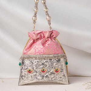 May include: A drawstring pouch bag featuring a pink fabric upper section with a floral pattern and a metallic silver lower section with intricate embossed scrollwork, three small crescent-shaped accents with green and red stones, and a beaded handle.