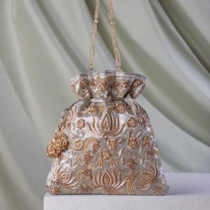 Puede incluir: Un bolso beige con cordón y bordado floral dorado. El bolso presenta un asa de cuentas y una pequeña borla. El diseño incluye costuras detalladas y una parte superior fruncida, creando una apariencia lujosa y ornamentada. Aproximadamente 20 cm de altura.