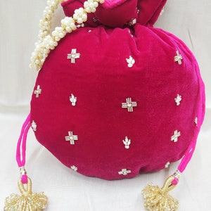 May include: A pink velvet drawstring bag with gold and white embellishments. The bag has a pearl beaded strap and gold tassel accents.