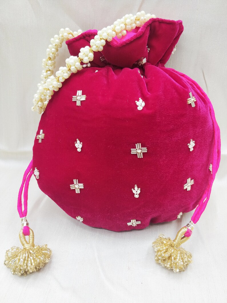 May include: A pink velvet drawstring bag with gold and white embellishments. The bag has a pearl beaded strap and gold tassel accents.