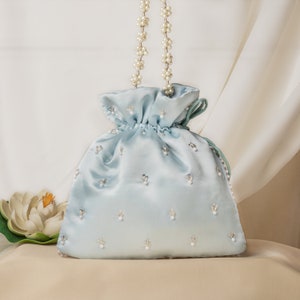 May include: A light blue satin drawstring bag with a pearl and silver embellishment. The bag has a pearl chain strap.