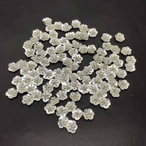 May include: A large quantity of silver flower-shaped sequins. The sequins are scattered on a black surface.