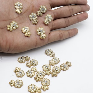 May include: A collection of gold-colored flower-shaped embellishments with pearl and rhinestone accents. The embellishments are scattered on a white surface.