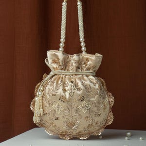 Puede incluir: Bolso de mano redondo de terciopelo beige con intrincados bordados florales y lentejuelas doradas. El bolso tiene asas con cuentas de perlas y un cierre de cordón. Una borla de perlas cuelga del lateral.