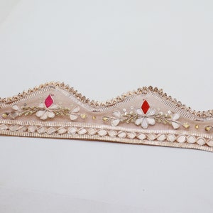 May include: Beige embroidered trim with a scalloped edge, featuring white floral designs, red and pink accents, and gold beads.