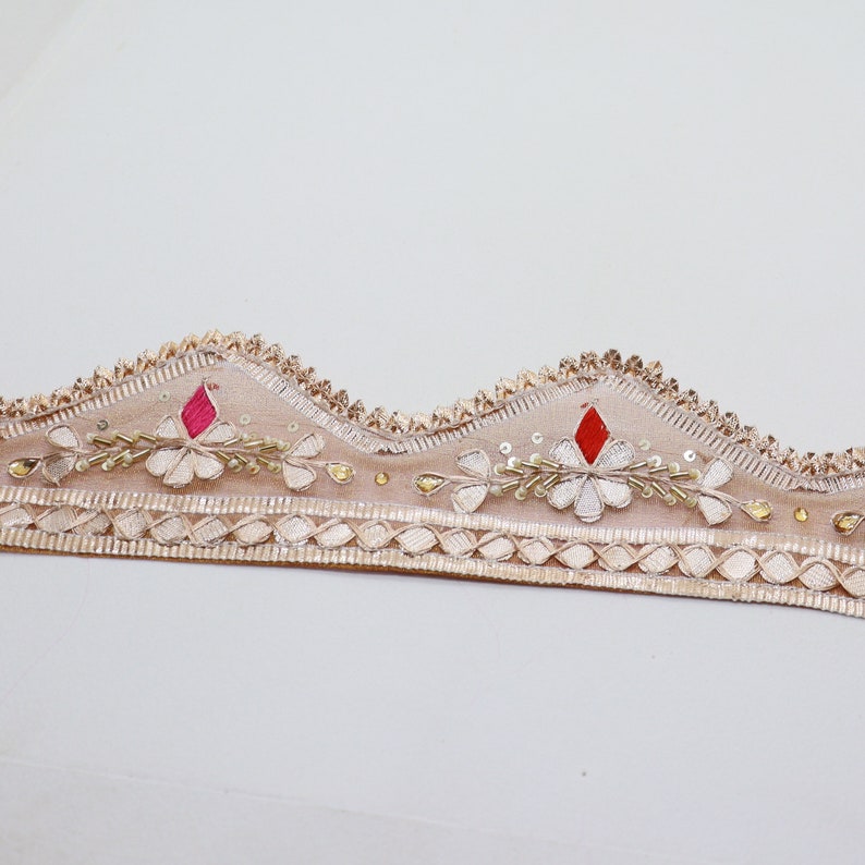 May include: Beige embroidered trim with a scalloped edge, featuring white floral designs, red and pink accents, and gold beads.