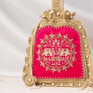 Könnte beinhalten: Ein fuchsiafarbener Samtbeutel mit Kordelzug und einer zentralen kreisförmigen Stickerei aus zwei Elefanten und Blumenmotiven in Goldfaden. Die Tasche ist mit aufwendigen goldenen Häkelarbeiten umrandet und mit kleinen weißen Perlen verziert.