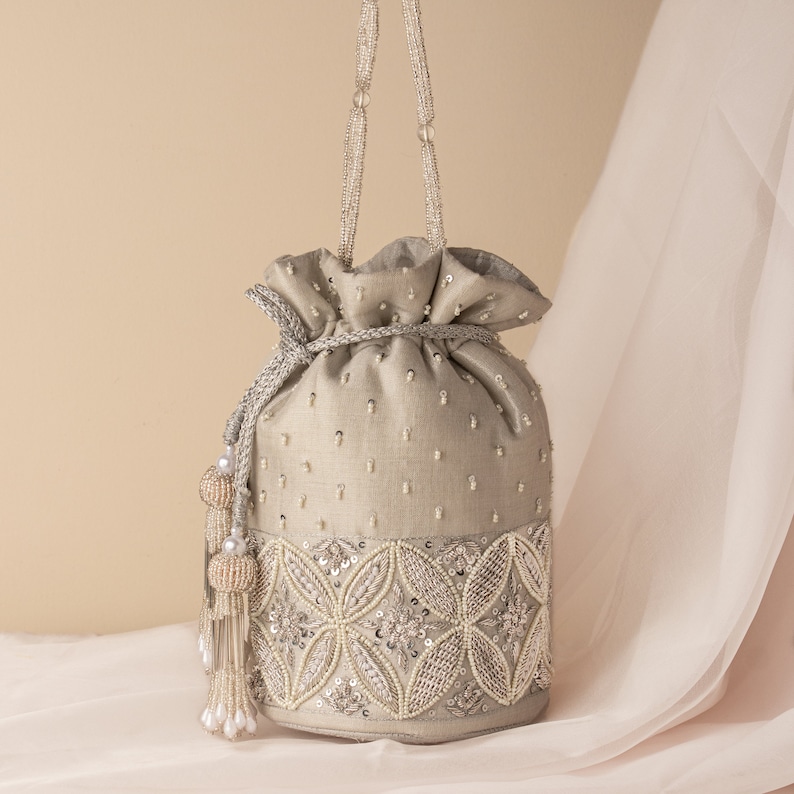 May include: A silver drawstring bag with a beaded design and a tassel. The bag has a silver chain strap and is decorated with pearls and sequins.