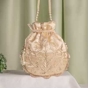 May include: A cream-colored satin drawstring bag with gold embroidery and pearl accents. The bag has a scalloped edge and a pearl chain strap.