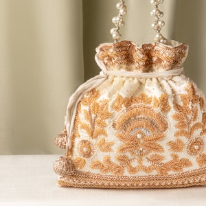 Könnte beinhalten: Eine weiße, perlenbesetzte Kordeltasche mit goldener Stickerei und einem Perlkettenhenkel.