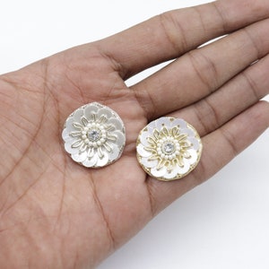 May include: Two white and silver floral embellishments with a central rhinestone and pearl accents. The circular design features petal-like sequins and a metallic silver or gold border. These decorative pieces are ideal for craft projects.