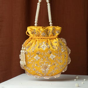May include: A yellow satin drawstring bag with intricate floral embroidery and embellishments. The bag features a pearl beaded handle and a tassel, with a scalloped edge. The bag is sitting on a white surface with a brown background.