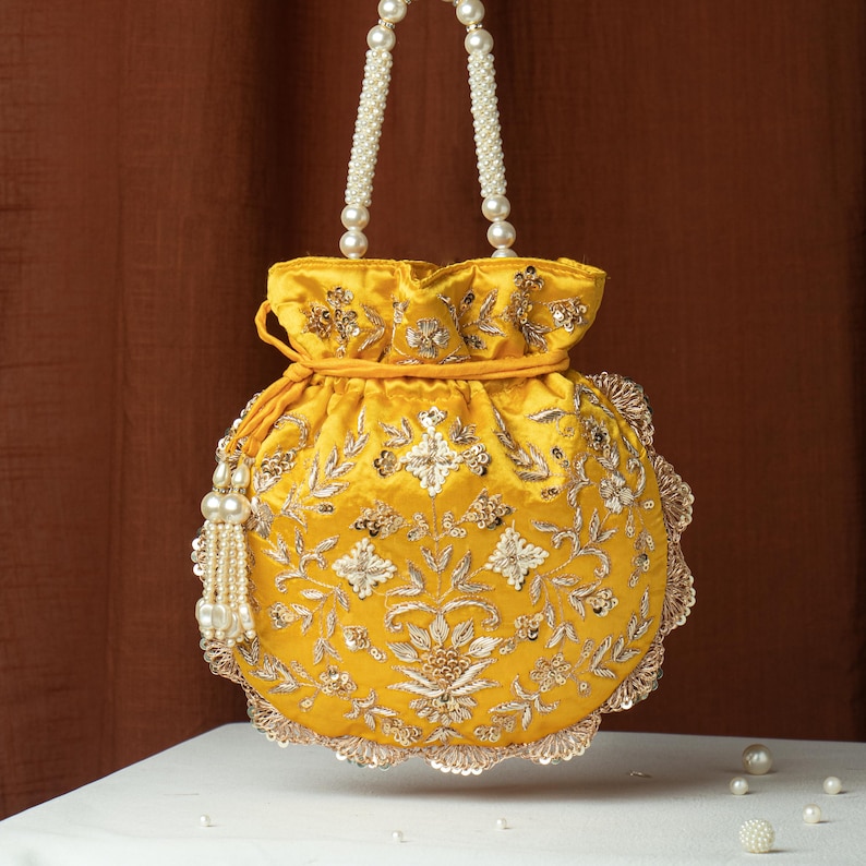 May include: A yellow satin drawstring bag with intricate floral embroidery and embellishments. The bag features a pearl beaded handle and a tassel, with a scalloped edge. The bag is sitting on a white surface with a brown background.