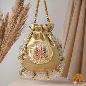 May include: Gold drawstring bag with intricate embroidery. The bag features a central circular design with colorful accents and ornate detailing. The handles are made of gold beads, and the edges are trimmed with lace and small gold bells.