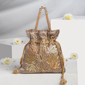 Könnte beinhalten: Ein goldfarbener Beutel mit aufwendiger floraler Stickerei. Die Tasche hat einen Perlenhenkel und Quasten. Das Design umfasst gold-, silber- und bronzefarbene Fäden, die ein strukturiertes, verziertes Aussehen erzeugen. Perlen und weiße Blumen sind um die Tasche verstreut.