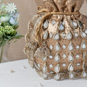 May include: A gold drawstring bag with detailed embroidery and mirror embellishments. The bag has teardrop-shaped mirror accents and a gold cord closure. A decorative tassel hangs from the side. The bag is on a white surface, with a bouquet of flowers in the background.