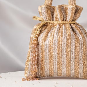 May include: A gold and silver drawstring bag with a vertical striped pattern. The bag has a gold cord closure and a decorative tassel with gold and clear beads. The bag is made of a shiny, textured fabric.