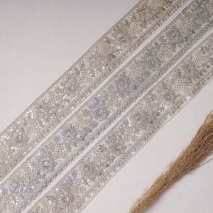 May include: A wide strip of sheer beige fabric trim featuring intricate silver metallic embroidery, floral patterns, and small clear sequins. The decorative border displays a repeating botanical design with dense stitching and shimmering accents.
