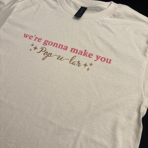May include: White t-shirt with the text "we're gonna make you Pop-u-lar" in pink lettering with gold glitter stars.