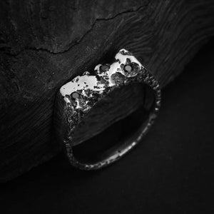 Peut inclure: Une bague en argent avec une surface texturée et rugueuse et trois pierres précieuses noires. La bague est sur un fond sombre et texturé.