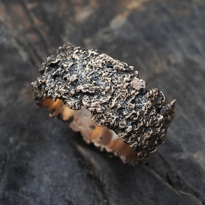 Op de afbeelding: Een brede, getextureerde bronzen ring met een ruw, organisch oppervlak. De ring is gemaakt van metaal en heeft een donkerbruine kleur.