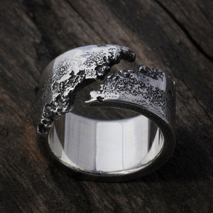 Puede incluir: Un anillo de plata con un diseño texturizado y rugoso. El anillo presenta un patrón dividido en forma de ola en la parte superior, creando una estética orgánica y moderna. La banda es lisa y pulida.