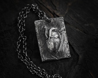 Op de afbeelding: Een zilveren ketting met een rechthoekige hanger met een gedetailleerd anatomisch hartontwerp. De hanger heeft een getextureerd oppervlak en zit aan een zwarte ketting.