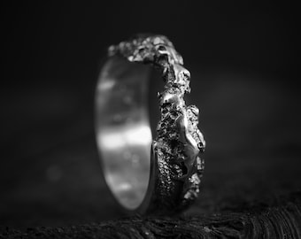 Può includere: Un anello d'argento con un design testurizzato e organico. L'anello è in metallo e ha una superficie ruvida e irregolare.