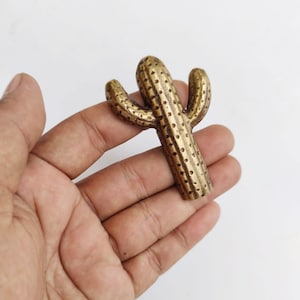 May include: A gold-colored metal cactus figurine with a textured surface. The cactus has three arms and is held in a hand.